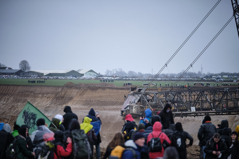 People stand next to the open pit mine looking towards Lützerath which is surrounded by police. 