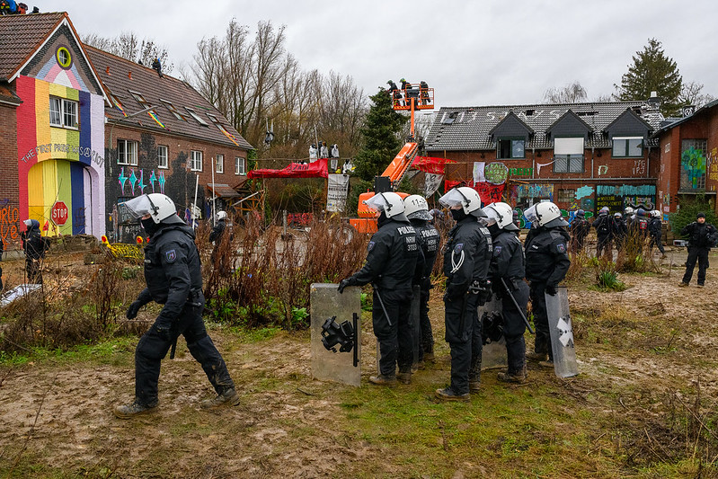 Riot police stand in from of a building, which has been painted with a pride flag. Activists sit on the roof of the building. In the background there are further buildings and heavy machinery.