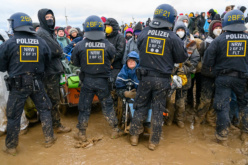 Four police people stand with the back to the photagrapher. A colorful crowd of protesters faces them, among the first row there is one person with wheelchair. The ground is very muddy. In the distance there are wind power plants.